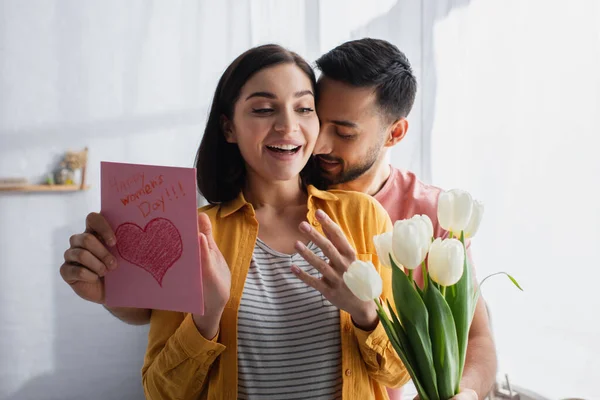 Junger Mann umarmt aufgeregte Freundin und überreicht Blumenstrauß mit Grußkarte in Küche — Stockfoto