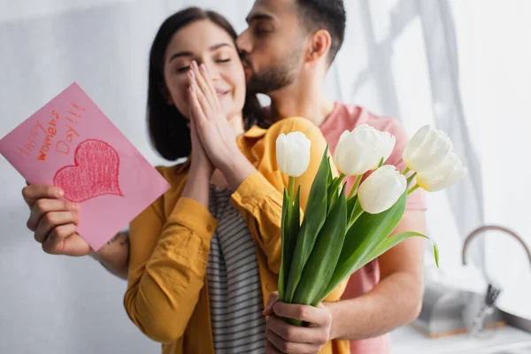 Young man kissing girlfriend and presenting bouquet of flowers with greeting card in kitchen — Stock Photo