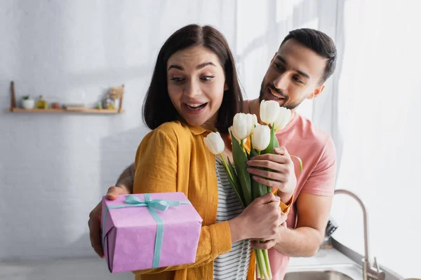 Junger Mann umarmt aufgeregte Freundin und präsentiert Blumenstrauß und Geschenkbox in Küche — Stockfoto