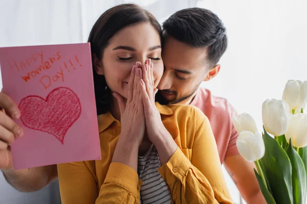 Junger Mann umarmt Freundin und überreicht Blumenstrauß und Grußkarte in Küche — Stockfoto