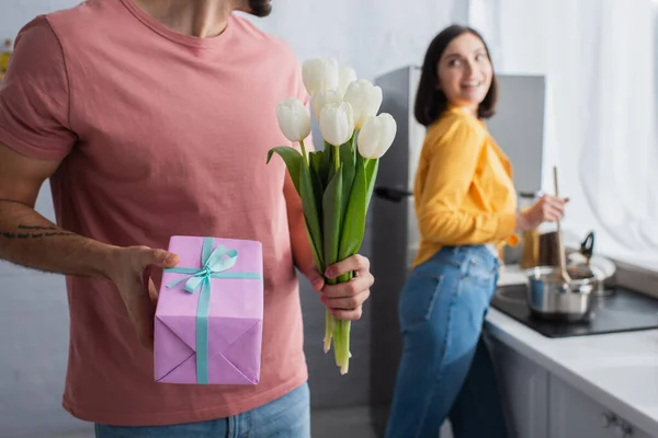 Jeune homme tenant bouquet de fleurs et boîte cadeau près de petite amie floue dans la cuisine — Photo de stock