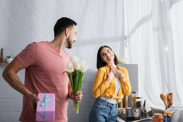 Smiling young man holding bouquet of flowers and gift box near girlfriend with hands on breast in kitchen — Stock Photo