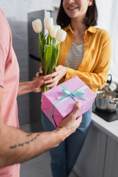 Partial view of young man presenting bouquet of flowers and gift box to girlfriend in kitchen — Stock Photo