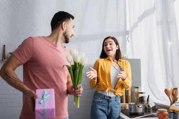 Animado jovem mulher com as mãos estendidas de pé perto do homem com buquê de flores e caixa de presente na cozinha — Fotografia de Stock