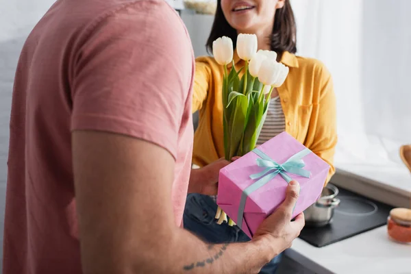 Частковий вид молодого чоловіка, що представляє букет квітів і подарункову коробку подрузі на кухні — стокове фото