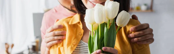 Teilansicht des jungen Mannes umarmt Freundin mit Blumenstrauß in Küche, Banner — Stockfoto