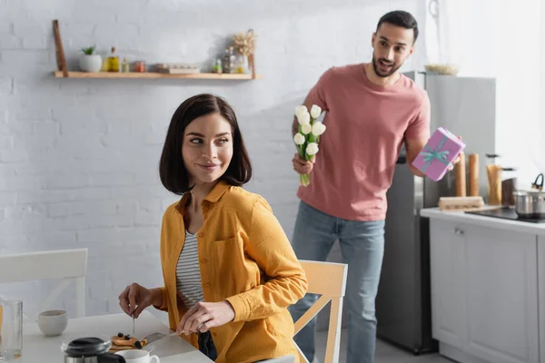 Sonriente mujer joven desayunando cerca borrosa novio con ramo de flores y caja de regalo en la cocina - foto de stock