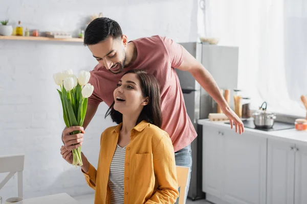 Lächelnder junger Mann überreicht aufgeregten Freundin in Küche Blumenstrauß mit offenem Mund — Stockfoto