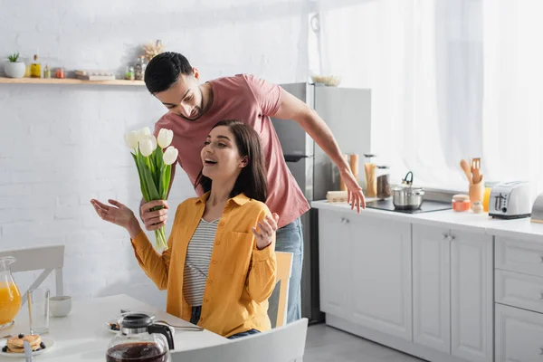 Jovem sorridente apresentando buquê de flores para namorada excitada com boca aberta e mãos estendidas na cozinha — Fotografia de Stock