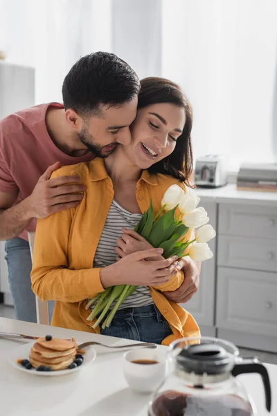 Sonriente joven abrazando novia con ramo de flores en la cocina - foto de stock