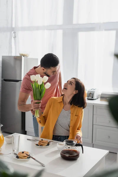 Jovem positivo apresentando buquê de flores para namorada excitada com boca aberta na cozinha — Fotografia de Stock