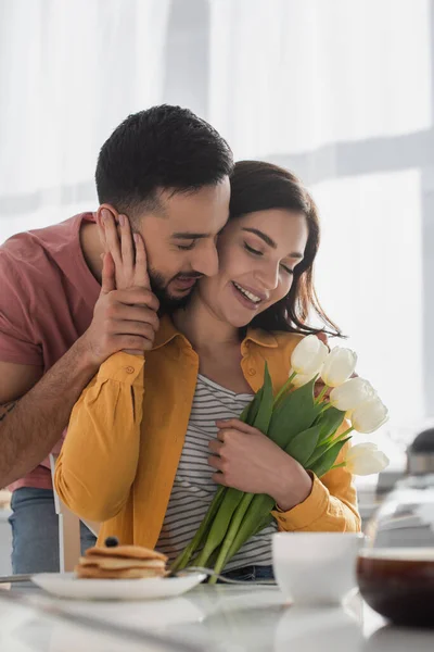 Young man hugging girlfriend with bouquet of flowers and closed eyes in kitchen — Stock Photo