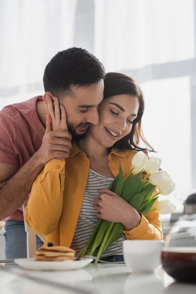 Joven abrazando novia con ramo de flores y ojos cerrados en la cocina - foto de stock