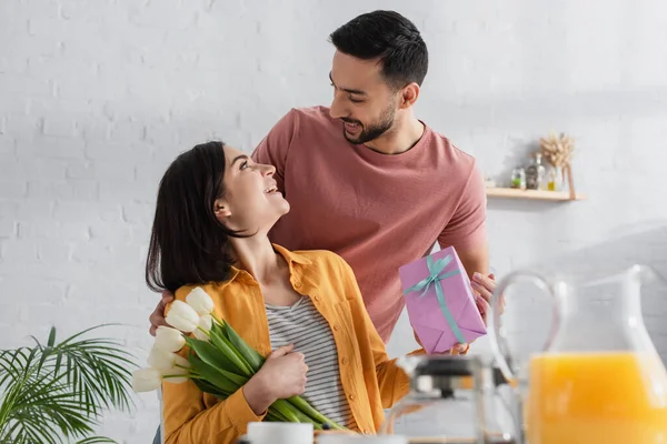 Positif jeune homme présentant boîte cadeau à petite amie avec bouquet de fleurs dans la cuisine — Photo de stock