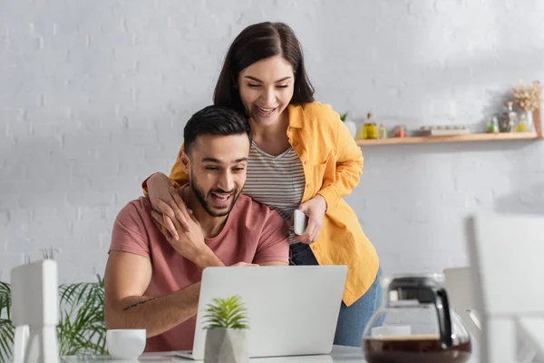 Sonriente mujer joven sosteniendo el teléfono celular y tocando las manos con el novio que trabaja con el ordenador portátil en casa - foto de stock