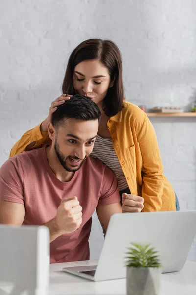 Junge Frau küsst Freund und schaut zu Hause mit Siegergesten auf Laptop — Stockfoto