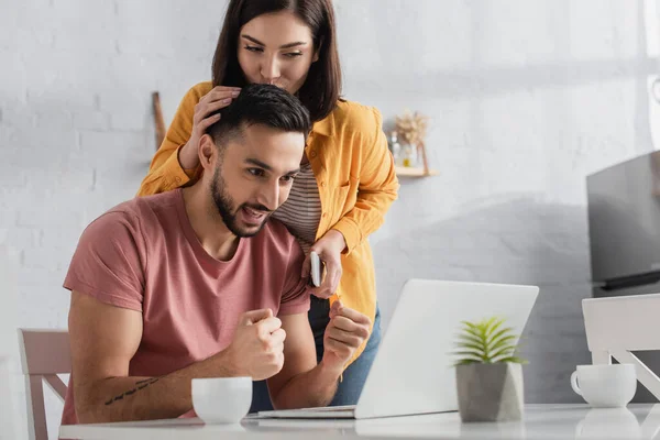 Mujer joven sosteniendo el teléfono celular y besando novio mirando a la computadora portátil con gestos de ganar en casa - foto de stock