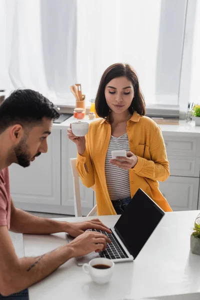 Sorridente giovane donna che beve caffè e messaggistica sul cellulare vicino al fidanzato che lavora con il computer portatile in cucina — Foto stock