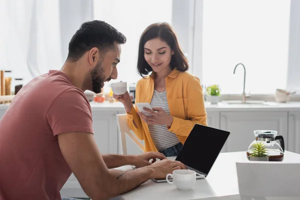 Sorridente giovane donna che beve caffè e messaggistica sul cellulare vicino al fidanzato che lavora con il computer portatile in cucina — Foto stock