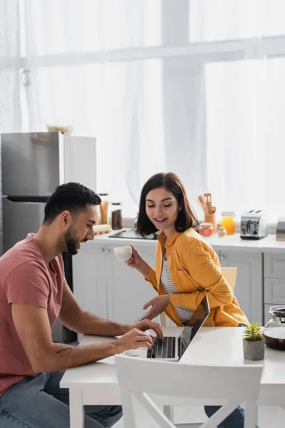 Lächelnde junge Frau trinkt Kaffee in der Nähe ihres Freundes, der in der Küche mit Laptop arbeitet — Stockfoto