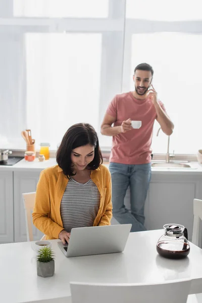 Улыбающаяся молодая женщина, работающая с ноутбуком рядом с размытым бойфрендом, говорящая по мобильному телефону на кухне — стоковое фото