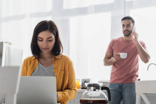 Giovane donna che lavora con il computer portatile vicino fidanzato offuscata parlando sul cellulare in cucina — Stock Photo