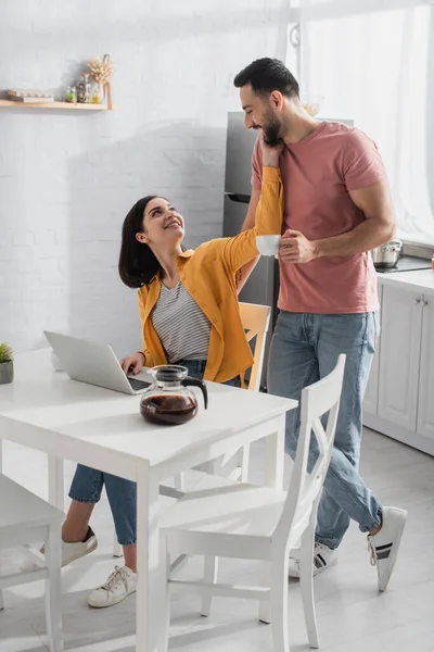 Smiling young man standing cup of coffee near girlfriend with laptop in kitchen — Stock Photo