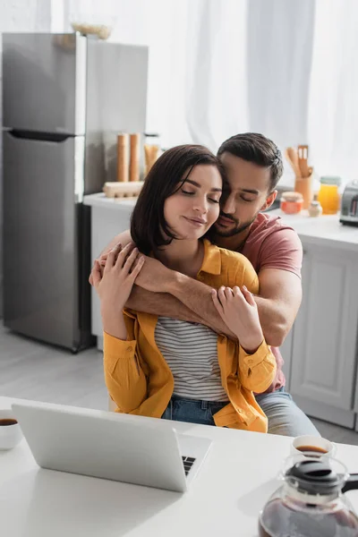 Smiling young man with closed eyes hugging girlfriend with laptop in kitchen — Stock Photo
