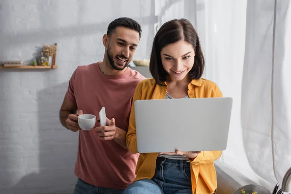 Smiling young man standing with cellphone and coffee cup near girlfriend sitting with laptop at home — Stock Photo