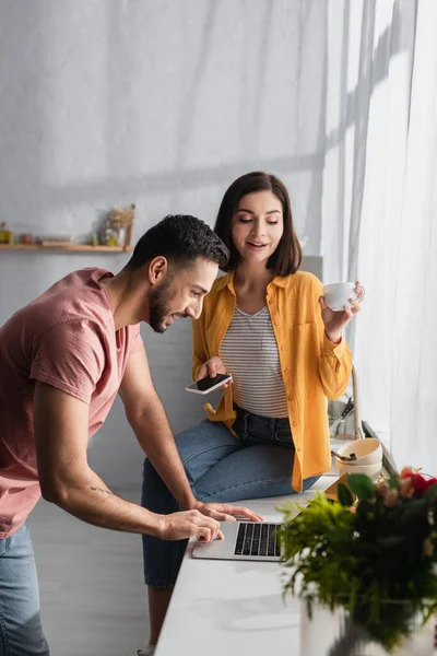 Souriant jeune femme assise avec téléphone portable et tasse de café près du petit ami avec ordinateur portable à la maison — Photo de stock