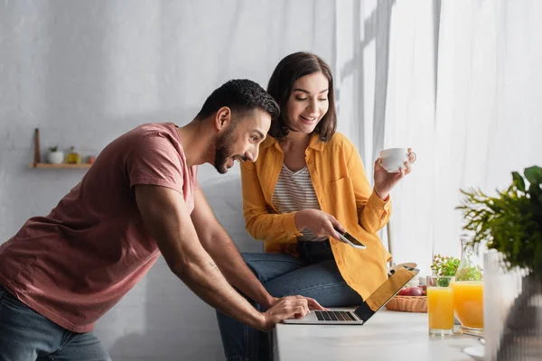 Souriant jeune femme assise avec téléphone portable et tasse de café près du petit ami avec ordinateur portable à la maison — Photo de stock