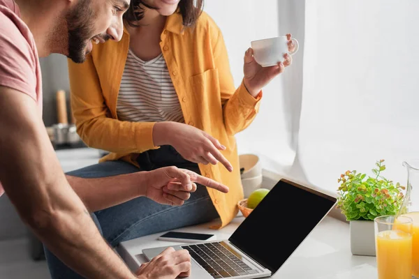 Vista parcial de pareja joven señalando con los dedos a la computadora portátil en casa - foto de stock