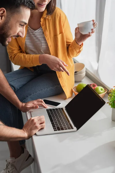 Mujer joven señalando con el dedo a la computadora portátil cerca de novio en casa - foto de stock