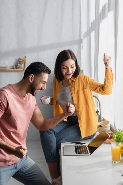 Excité jeune couple regardant ordinateur portable avec des gestes de victoire à la maison — Photo de stock