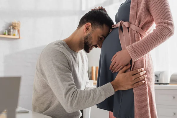 Feliz joven tocando vientre de la mujer embarazada con la cabeza en la cocina - foto de stock