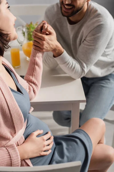 Vista parcial del joven sentado a la mesa y cogido de la mano de la mujer embarazada en la cocina - foto de stock