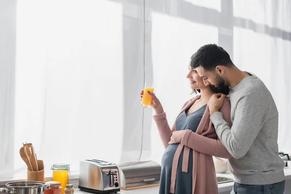 Joven con los ojos cerrados abrazando suavemente a la mujer embarazada con jugo de naranja en la cocina - foto de stock