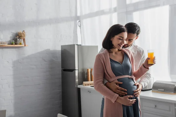 Giovane uomo ad occhi chiusi che abbraccia dolcemente una donna incinta con succo d'arancia in cucina — Foto stock