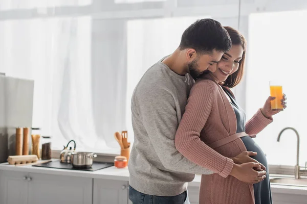 Giovane uomo ad occhi chiusi che abbraccia dolcemente una donna incinta con succo d'arancia in cucina — Foto stock