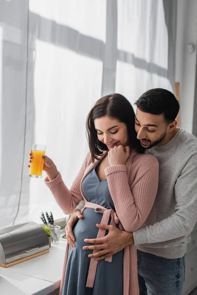 Excited young man gently hugging pregnant woman with orange juice in kitchen — Stock Photo