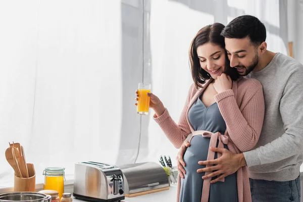 Giovane uomo sorridente che abbraccia dolcemente una donna incinta con succo d'arancia in cucina — Foto stock