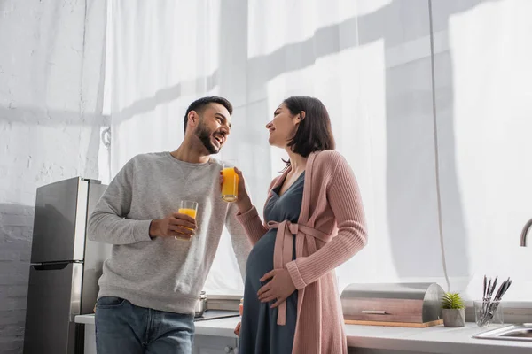 Счастливый молодой человек и беременная женщина стоят с апельсиновым соком на кухне — стоковое фото