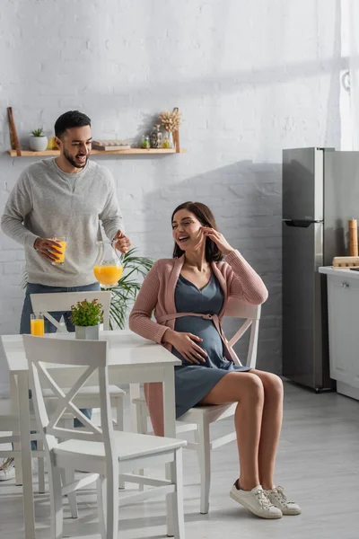 Sonriente joven de pie con frasco y vaso de jugo de naranja cerca de la mujer embarazada en la cocina - foto de stock