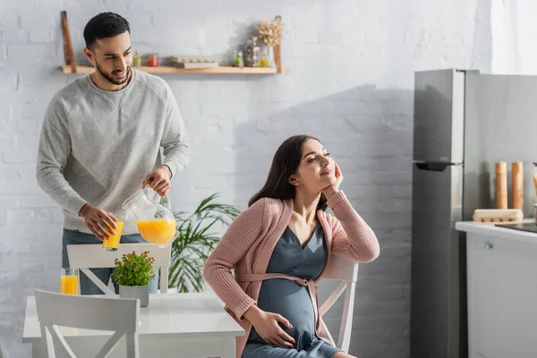 Positive young man pouring orange juice from jar to glass near pregnant woman in kitchen — Stock Photo