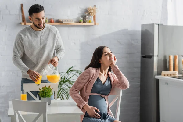 Hombre joven positivo vertiendo jugo de naranja del frasco al vidrio cerca de la mujer embarazada en la cocina - foto de stock