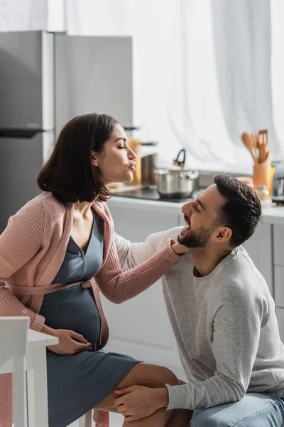 Giovane uomo sorridente seduto vicino a una donna incinta con le labbra indolenzite in cucina — Foto stock