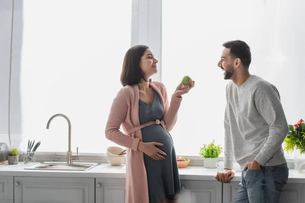 Усміхнений молодий чоловік стоїть біля вагітної жінки з яблуком на кухні — стокове фото