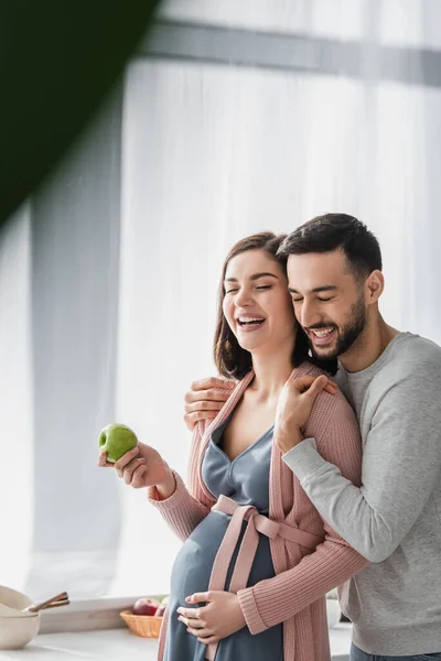 Sonriente joven con los ojos cerrados abrazando a la mujer embarazada con manzana en la cocina - foto de stock