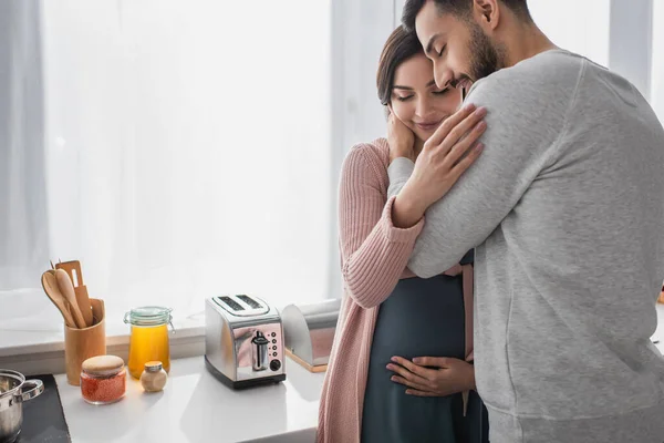 Sorridente jovem com olhos fechados abraçando mulher grávida na cozinha — Fotografia de Stock