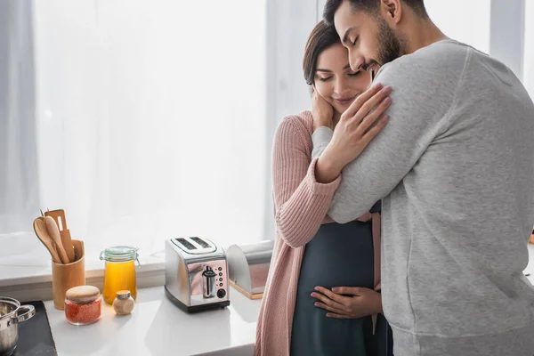 Sonriente joven con los ojos cerrados abrazando a la mujer embarazada en la cocina - foto de stock